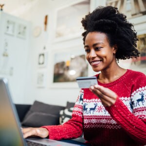 A black woman in a holiday sweater holds a credit card in her hand while using her laptop to shop online, meaning she understands how to know if a website is safe to purchase from. 