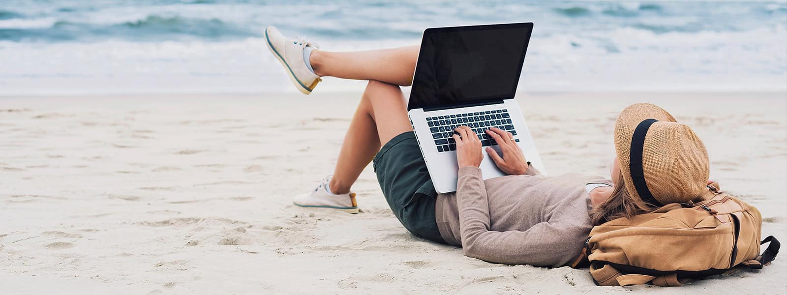 woman wearing a hat, laying on the beach with her laptop and backpack.