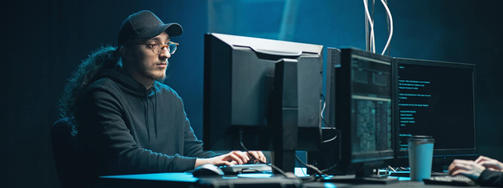 A man with glasses, a black ball cap, and a black sweatshirt sits in a dark room coding at a desktop computer