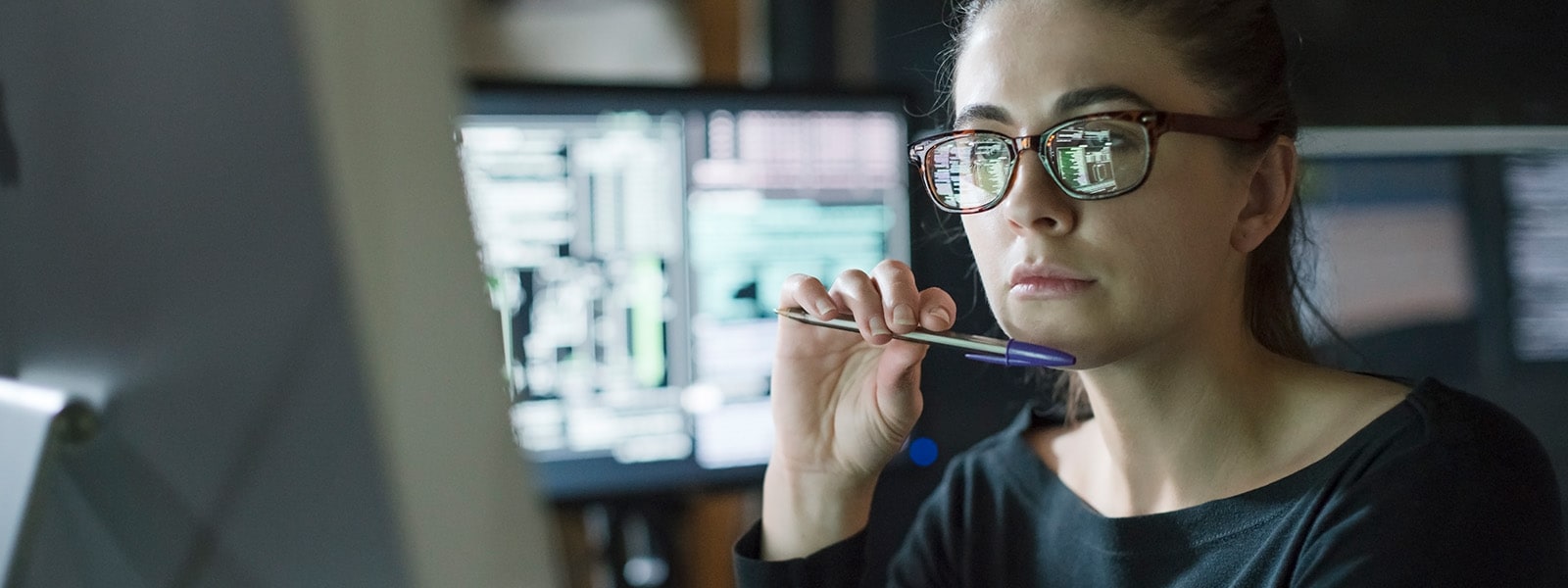 A contemplative woman in glasses holds a pen and gazes at a computer monitor, suggesting she is checking for spyware. 