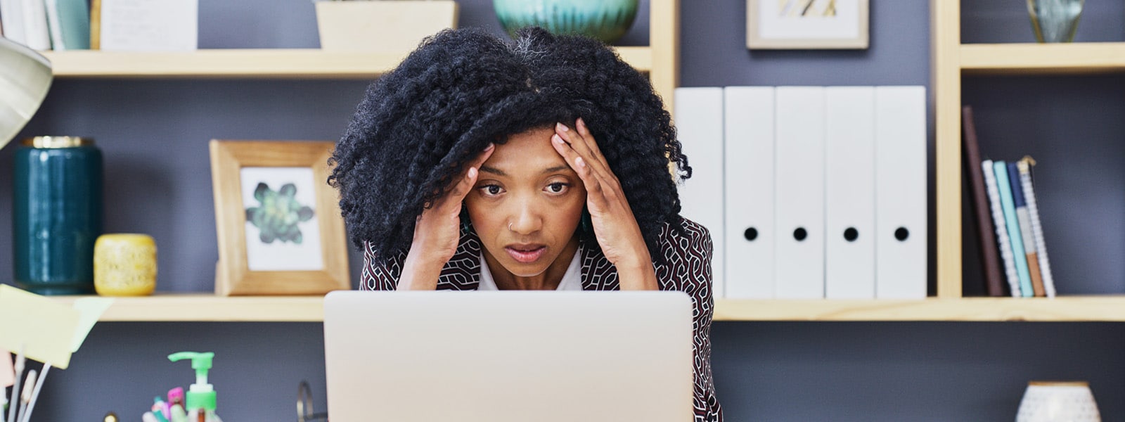 a black woman in an office stares at her laptop with hands pressed to her forehead, indicating she might be experiencing some type of malware