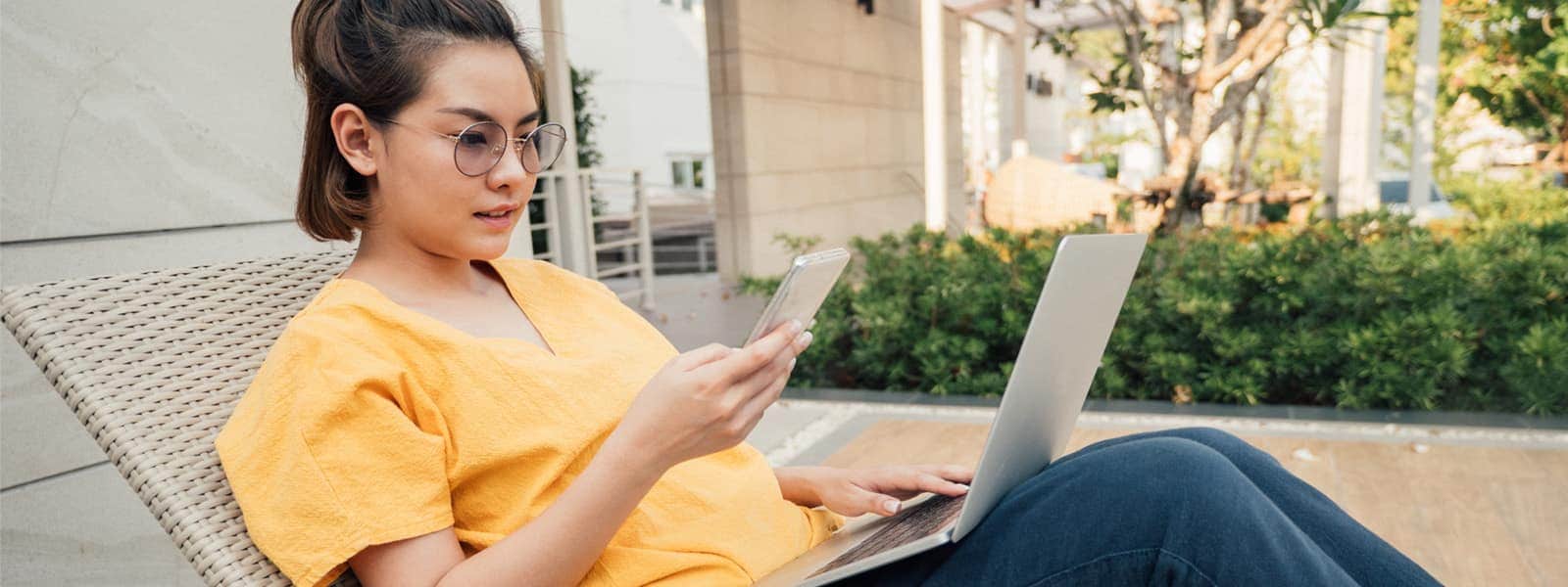 an Asian woman in an orange blouse lounges outside with a laptop in her lap and a cellphone in her hand, indicating she might be investing in cryptocurrency and also experienced a cryptocurrency scam