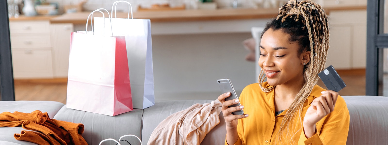 a black woman in a yellow shirt sits on her couch, surrounded by shopping bags and with a credit card in hand one hand and a phone in the other, indicating she is online shopping